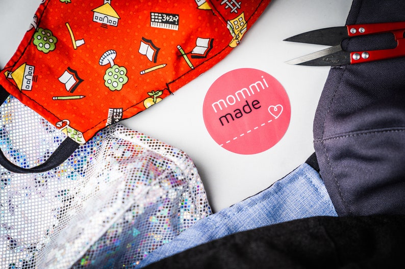 May include: A close-up of a variety of fabrics, including a red fabric with a school-themed pattern, a silver sequined fabric, a blue denim fabric, and a black fabric. The fabrics are arranged on a white surface with a pink circle sticker that says "mommi made" with a heart.