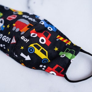 May include: A black face mask with a colorful car pattern. The mask features various vehicles, road signs, and the words "GO GO!" in white. The mask has black elastic ear loops.
