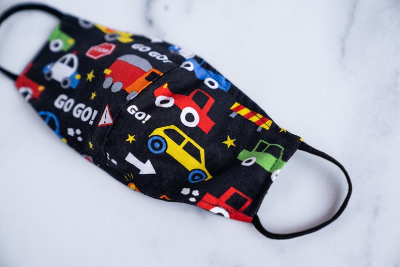 May include: A black face mask with a colorful car pattern. The mask features various vehicles, road signs, and the words "GO GO!" in white. The mask has black elastic ear loops.