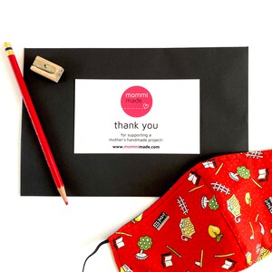 May include: A red pencil and pencil sharpener are next to a black card with a white card that says "mommi made" and "thank you". A red face mask with a school-themed pattern is also in the image.