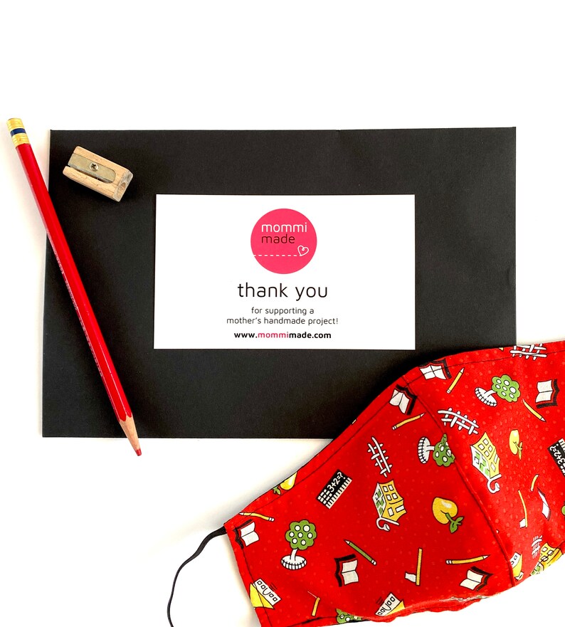 May include: A red pencil and pencil sharpener are next to a black card with a white card that says "mommi made" and "thank you". A red face mask with a school-themed pattern is also in the image.