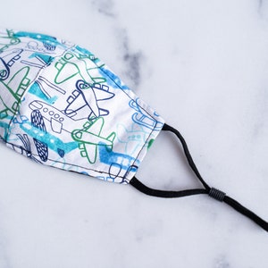 May include: A white face mask with a pattern of blue and green airplanes. The mask has black adjustable ear loops and is made of fabric. The airplanes are in a cartoon style.