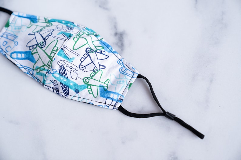May include: A white face mask with a pattern of blue and green airplanes. The mask has black adjustable ear loops and is made of fabric. The airplanes are in a cartoon style.