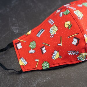 May include: Red fabric face mask with a repeating pattern of school-themed illustrations. The pattern includes books, pencils, trees, and buildings. The mask has a black elastic band for securing it around the ears.