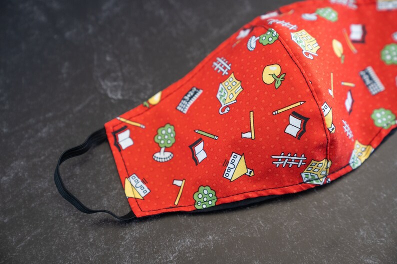 May include: Red fabric face mask with a repeating pattern of school-themed illustrations. The pattern includes books, pencils, trees, and buildings. The mask has a black elastic band for securing it around the ears.