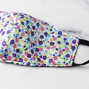 May include: A floral face mask with a white background. The mask features a pattern of small purple, pink, and blue flowers with green stems and leaves. It has a black elastic band for securing it around the ears.