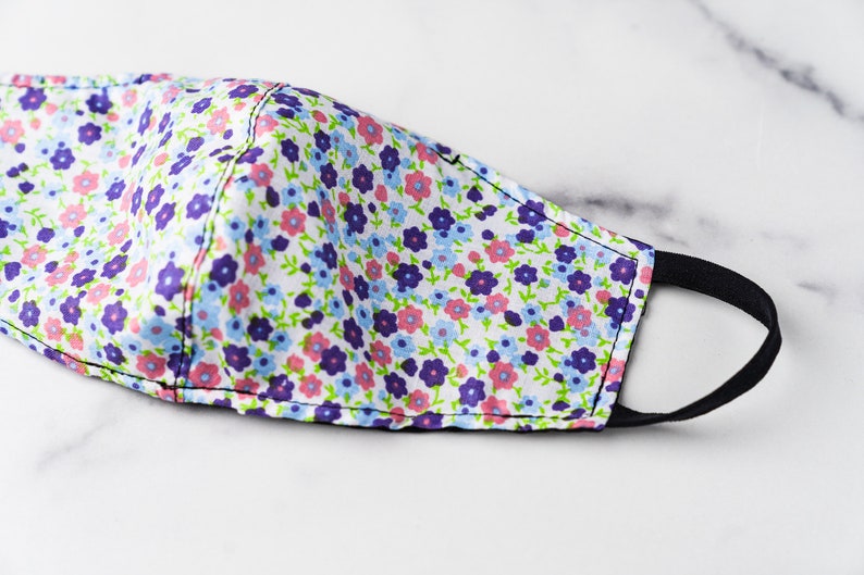 May include: A floral face mask with a white background. The mask features a pattern of small purple, pink, and blue flowers with green stems and leaves. It has a black elastic band for securing it around the ears.