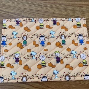 May include: A rectangular quilted fabric mat featuring a repeating pattern of Peanuts characters including Charlie Brown, Snoopy, Lucy, and Linus interspersed with orange pumpkins and brown autumn leaves on a light peach background.