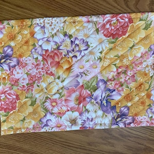 May include: A rectangular quilted placemat featuring a dense floral pattern with various blossoms in shades of yellow, pink, purple, and white. The fabric displays a mix of daffodils, daisies, and other flowers with diagonal stitching lines.