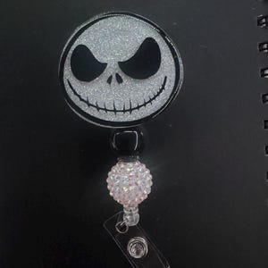 May include: A round, glittery badge reel featuring Jack Skellington's face from The Nightmare Before Christmas. The design is black and white with a silver glitter background. The reel has a black bead, a pink rhinestone bead, and a clear plastic clip.