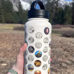 May include: White water bottle with a black handle, covered in circular stickers featuring various landscape illustrations. The bottle is held outdoors with a mountain backdrop. The stickers depict scenes of nature and landmarks.