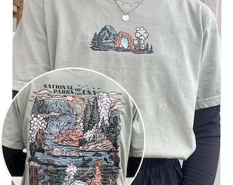 May include: A light green t-shirt with a graphic print of a national park scene. The print features a mountain range, a natural arch, and a river. The text "National Parks of the USA" is printed on the shirt.