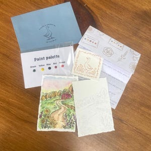 Puede incluir: Un kit de pintura de acuarela que incluye un sobre azul con una ilustración de un ganso, una tarjeta de paleta con cinco puntos de pintura etiquetados Green, Yellow, Blue, Red, Orange, una pequeña impresión de ganso y dos hojas de papel de acuarela.