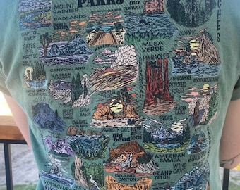 May include: A green t-shirt featuring a map of the United States depicting national parks. The map is illustrated with various landmarks and features of each park, including mountains, trees, and water. The text "National Parks of the USA" is printed in black at the top of the map.