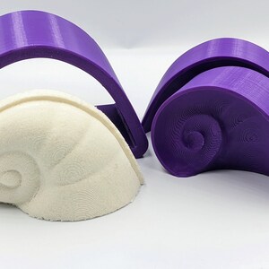 May include: Two purple 3D printed molds for making a seashell shape. One mold is a single piece, the other is in two pieces. A white seashell is sitting in the single piece mold.