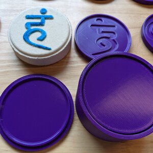 May include: A set of five purple round coasters with a raised design. One coaster has the Sanskrit symbol "Om" in the center. Another coaster has the Sanskrit symbol "Aum" in the center. The remaining three coasters are plain.