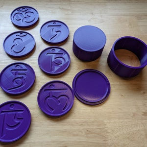 May include: A set of nine purple 3D printed coasters with different symbols, including Om and other Sanskrit characters. The coasters are arranged on a wooden surface, along with two purple 3D printed containers.
