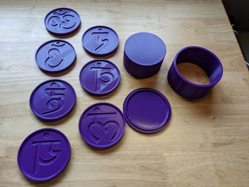 May include: A set of nine purple 3D printed coasters with different symbols, including Om and other Sanskrit characters. The coasters are arranged on a wooden surface, along with two purple 3D printed containers.