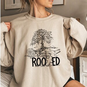 May include: A beige sweatshirt with a black graphic of a tree growing out of a book. The text "stay rooted" is printed below the tree.