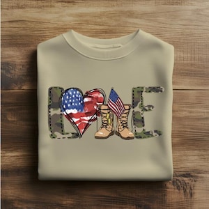 Könnte beinhalten: Ein beiges Sweatshirt mit Rundhalsausschnitt und einem Grafikdruck des Wortes LOVE. Das L und E haben ein grünes und braunes Tarnmuster. Das O ist ein Herz mit amerikanischem Flaggendesign, und das V wird durch zwei braune Stiefel ersetzt.