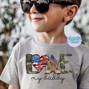 May include: A grey t-shirt featuring a graphic design of a heart with an American flag, camouflage, and boots. The text "Love my daddy" is below the graphic.