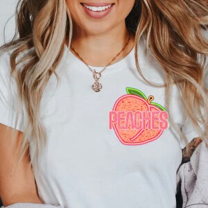 May include: White t-shirt featuring a peach graphic with the text "Peaches" in neon pink lettering.