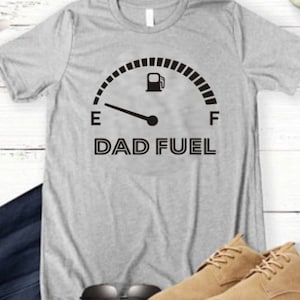Puede incluir: Una camiseta gris con un gráfico de indicador de combustible negro y el texto "DAD FUEL" impreso en ella.