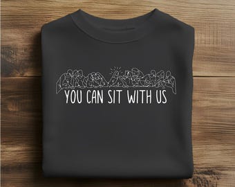 May include: A black crewneck sweatshirt with a white graphic of people sitting together, with the text "YOU CAN SIT WITH US" below the graphic. The sweatshirt is made of a soft material.