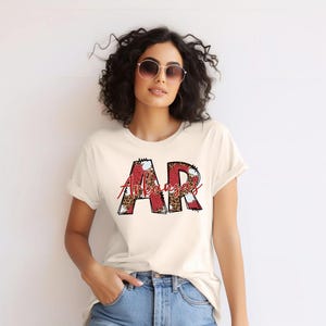 May include: A white t-shirt with a red and black graphic design of the letters "AR" with the word "Arkansas" written underneath.