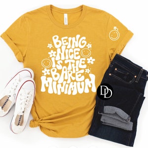May include: A mustard yellow t-shirt with the text "Being Nice Is The Bare Minimum" in white, with a flower and smiley face design. The shirt is laid flat on a white surface with a pair of white sneakers and blue jeans.