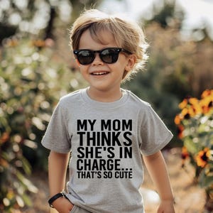 May include: A young child wearing a grey t-shirt with the text "My Mom Thinks She's In Charge... That's So Cute" printed in black. The child is wearing sunglasses and smiling.
