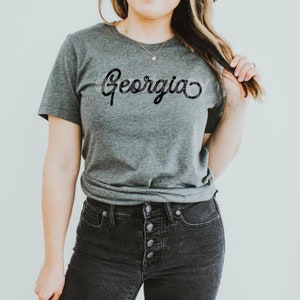 以下が含まれることがあります： ヘザーグレーのTシャツに、黒い筆記体で「ジョージア」と書かれています。