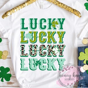 May include: White t-shirt with the words "Lucky" repeated four times in green, blue, and leopard print lettering. Each word has a shamrock next to it.