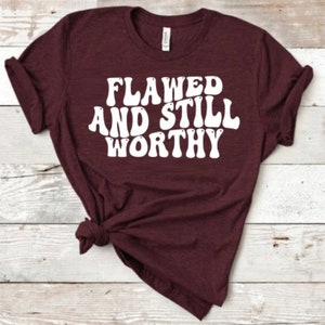 May include: A maroon t-shirt with the text "Flawed and Still Worthy" in white, retro-style lettering.