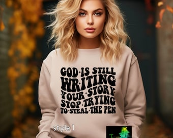 May include: A beige sweatshirt with a black graphic print that reads "God is still writing your story stop trying to steal the pen". The text is in a retro font style. The sweatshirt has a crew neck and long sleeves.