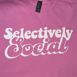 May include: Pink t-shirt with the text "Selectively Social" in white, retro-style lettering.