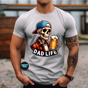May include: A grey t-shirt featuring a cartoon skeleton wearing a plaid shirt and a blue baseball cap. The skeleton is holding a glass of beer and the text "DAD LIFE" is printed below the image.