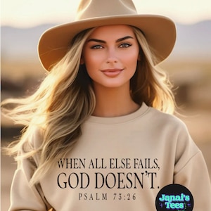 May include: A woman wearing a tan sweatshirt with the text "WHEN ALL ELSE FAILS, GOD DOESN'T. PSALM 73:26" and a tan hat. The text "Janai's Tees" is in a circle with a pink, blue, and white design.