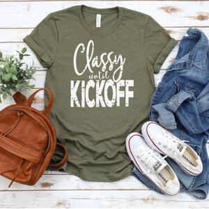 May include: A green t-shirt with the text "Classy until Kickoff" in white lettering. The shirt is laid out on a white wooden surface with a brown leather backpack, a pair of white trainers, and a blue denim jacket.