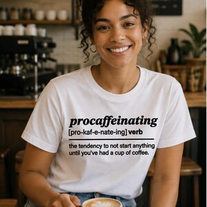 May include: A white t-shirt with the word "procaffeinating" printed in black, along with its pronunciation and definition. The person is holding a cup of coffee with latte art. The image is taken in a coffee shop.