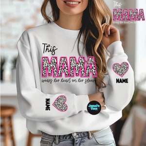 Könnte beinhalten: Weißer Pullover mit dem Text "This Mama wears her heart on her sleeve" in Pink und schwarzem Leopardenmuster. Der Pullover hat ein pinkfarbenes Herz mit einem Leopardenmuster auf dem Ärmel.