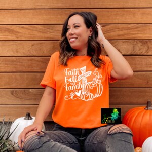 Könnte beinhalten: Eine Frau trägt ein orangefarbenes T-Shirt mit dem Text "Faith Fall and Family" und einem weißen Kreuz- und Kürbisdesign. Sie sitzt vor einer Holzwand mit Kürbissen.