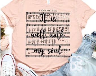 May include: A peach-colored short-sleeve t-shirt featuring black printed musical staff lines and lyrics from the hymn It Is Well with My Soul. The text It is well with my soul is written in a large black cursive script over the sheet music.