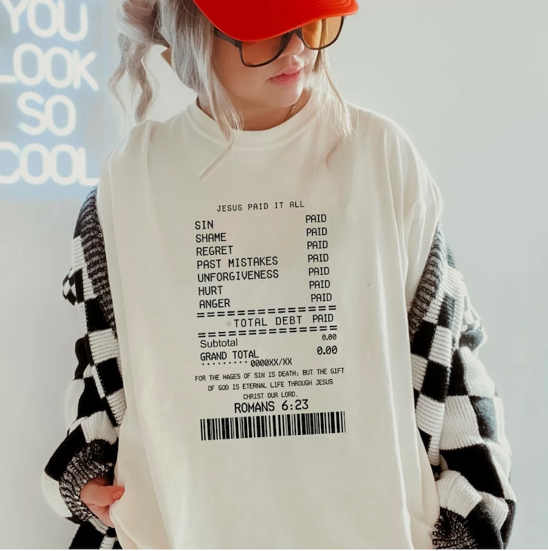 K&ouml;nnte beinhalten: Wei&szlig;es Sweatshirt mit einem schwarz-wei&szlig; karierten Muster am &Auml;rmel. Die Vorderseite des Sweatshirts zeigt eine Quittungsgrafik mit dem Text "Jesus Paid It All" und einer Liste von S&uuml;nden mit dem Wort "Paid" neben jeder. Die Quittung enth&auml;lt auch den Text "Total Debt Paid" und "R&ouml;mer 6:23".