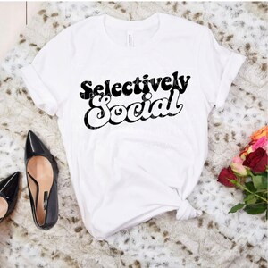 May include: White t-shirt with black text that reads "Selectively Social" in a retro font style.