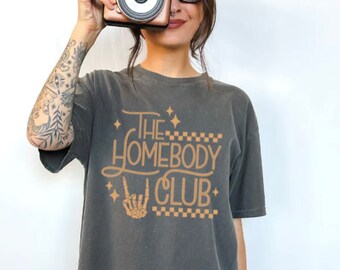 May include: A gray t-shirt with the text "The Homebody Club" in orange lettering. The text is stylized with a checkered pattern and a skeleton hand making a rock and roll hand gesture.