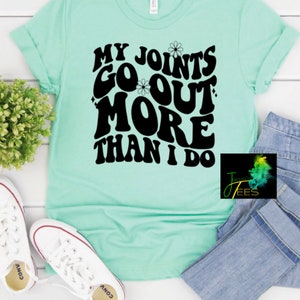 May include: A light green t-shirt with the text "My Joints Go Out More Than I Do" in black, stylized lettering. The text is surrounded by small white flower graphics.