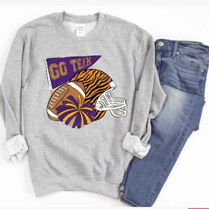 May include: Grey sweatshirt featuring a graphic of a football helmet, football, and a pennant that reads "GO TEAM". The helmet is striped with orange and black, and the football has purple and gold pom-poms.