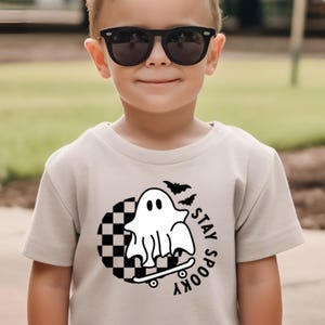 May include: A light grey t-shirt with a black and white chequered circle design. A ghost is skateboarding in the circle with the text "Stay Spooky" written vertically.