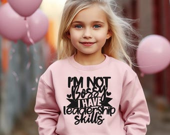 Pode incluir: Camisola rosa claro com o texto preto "I'M NOT Bossy I HAVE leadership skills". Uma menina loira usa a camisola. Balões cor-de-rosa são visíveis no fundo.